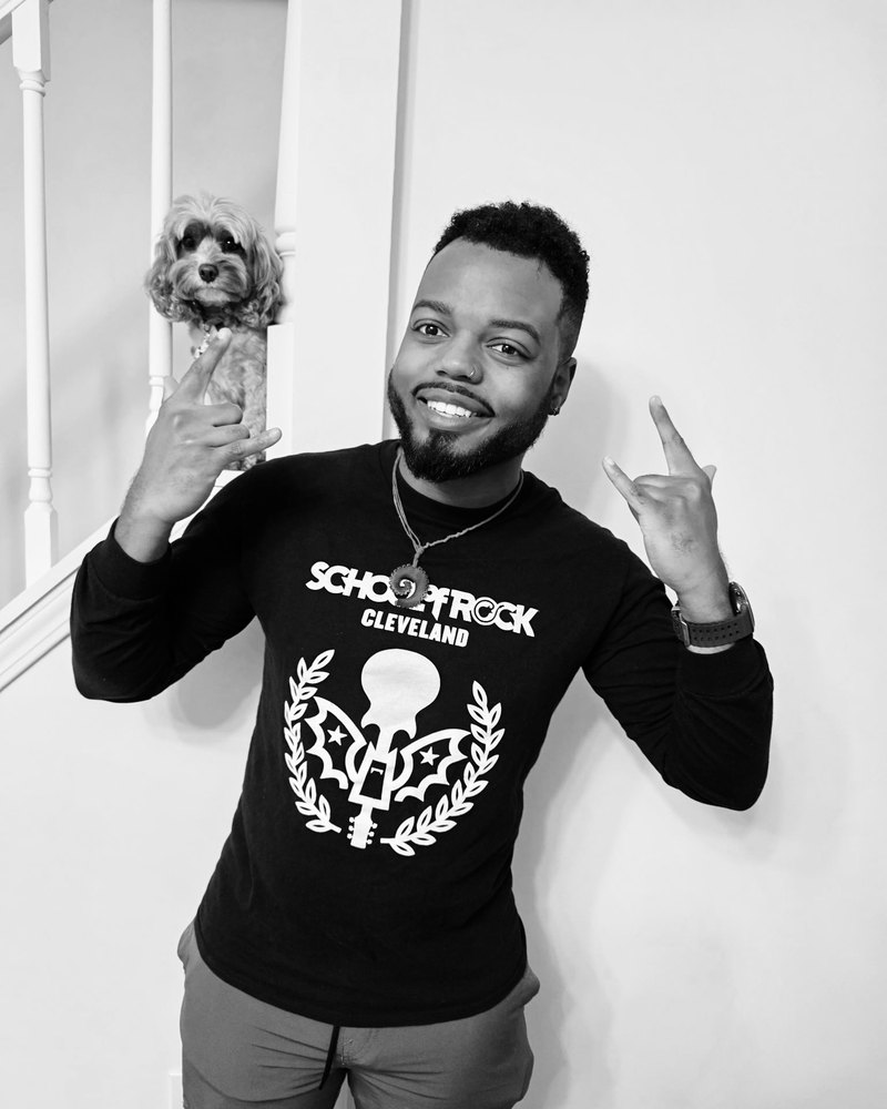 Black-and-white photo of Justis “The Poet” Daniels-Bezout in a School of Rock Cleveland shirt, making rock-on hand signs, with a small dog on the stair rail behind him